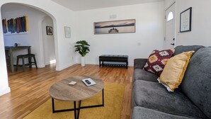 Uma sala de estar com um sofá cinzento, uma mesa de centro redonda, um tapete amarelo, um vaso de planta e um quadro na parede.