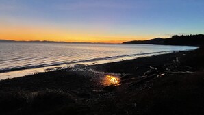 A beach at sunset with calm waters, a campfire, and distant hills.