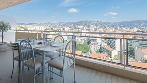A balcony with a table and chairs, overlooking a cityscape with buildings and hills. - 3-Br. apartment near downtown Cannes. Sea view, parking. (Cannes)