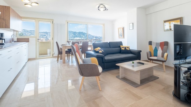 A modern living room with a sofa, coffee table, and TV stand. There is a dining area with a table and chairs, and a kitchen with a stove and sink. - 3-Br. apartment near downtown Cannes. Sea view, parking. (Cannes)