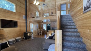Una cabaña de madera espaciosa con una zona de estar central, chimenea, televisor y una escalera que lleva a un nivel superior.