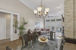 A spacious living area with a glass dining table, wicker chairs, a brown leather sofa, a chandelier, and a view to the outside.