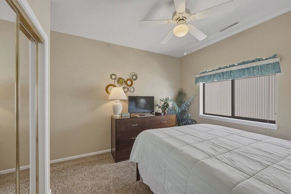 A bedroom with a bed, a dresser, a TV, a lamp, a ceiling fan, and a window with blinds.