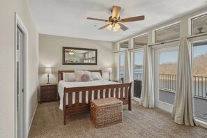 A bedroom with a large bed, a ceiling fan, a mirror, a window with curtains, and a balcony with a view.