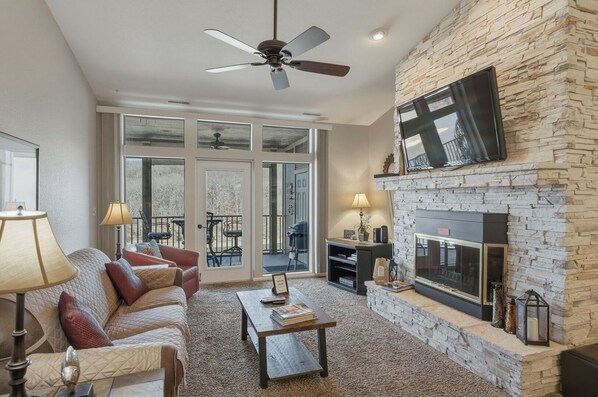 A living room with a stone fireplace, a ceiling fan, and a large TV mounted on the wall.