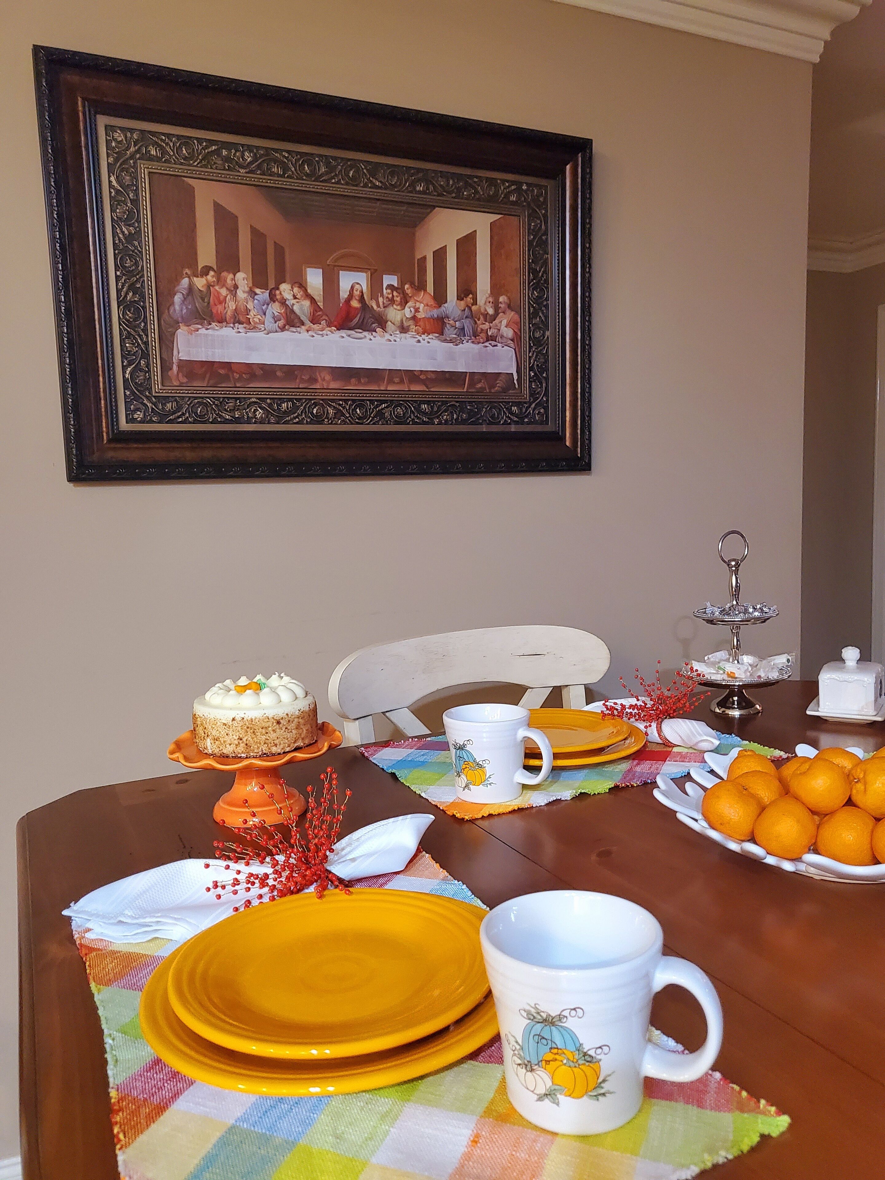 A dining table set with yellow plates, a white mug with a pumpkin design, a cake, and a plate of oranges.