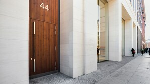 Fachada moderna de edificio con una puerta de madera grande marcada con el número 44, un primer piso limpio con baldosas y un paseo peatonal.