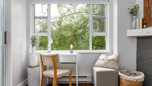 A corner room with a white desk, a chair, a small grey sofa, a wicker basket, and a large window with a view of trees.