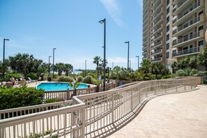 Zona de piscina en un resort, con una piscina azul cristalina, rodeada de palmeras y una valla blanca.