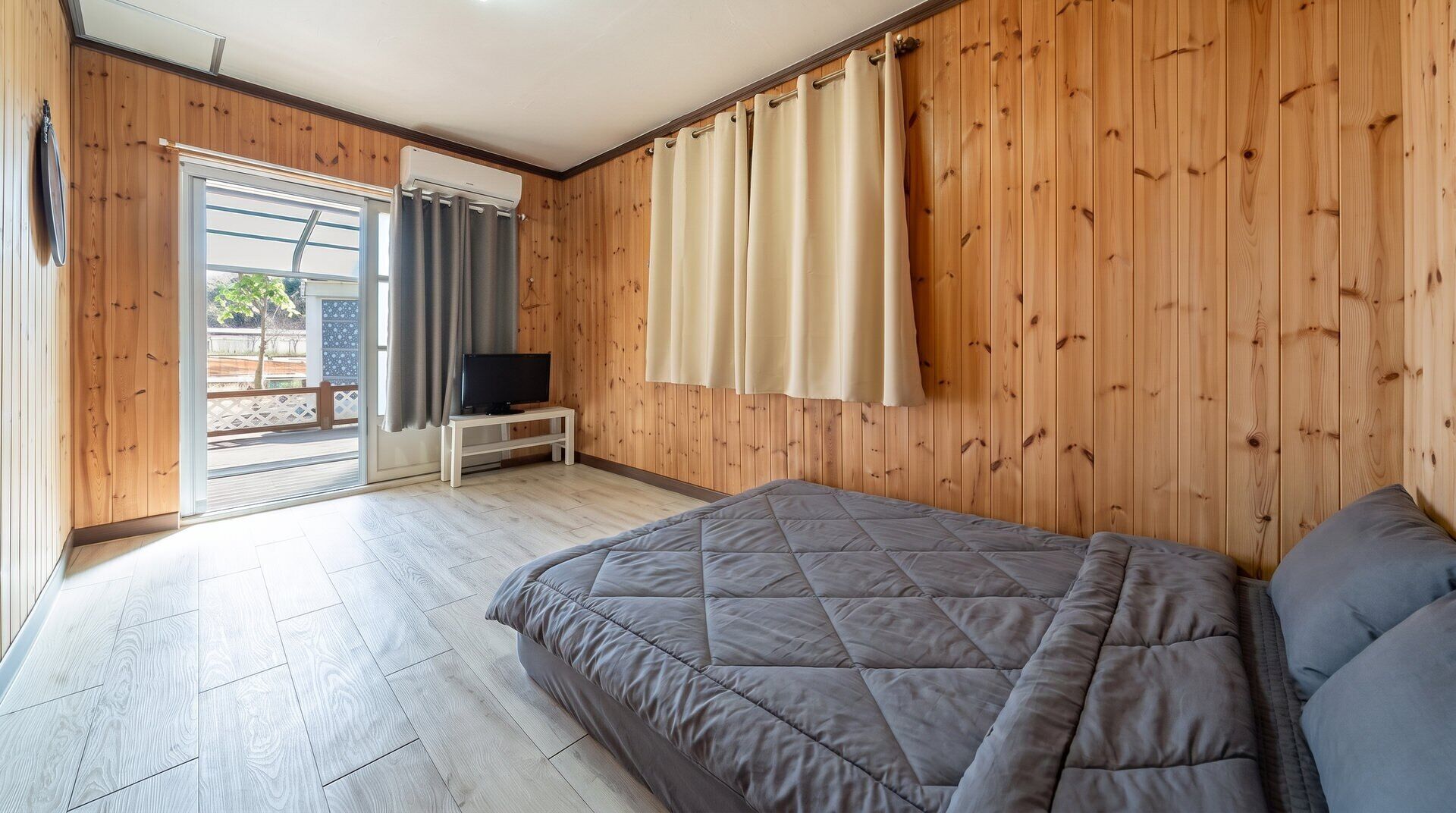 A bedroom with a bed, a television on a small table, wooden walls, and a sliding glass door leading to an outdoor area.