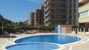 Una piscina rodeada de jardines con palmeras y un edificio de varios pisos al fondo.