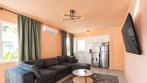 A compact living space with a sofa, a small round table, a kitchen area with a refrigerator and stove, and a mounted TV.