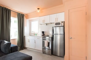 A compact kitchen with white cabinets, a stainless steel refrigerator, a microwave, and a stove. There is a window with curtains, a sink, and a countertop with a kettle.