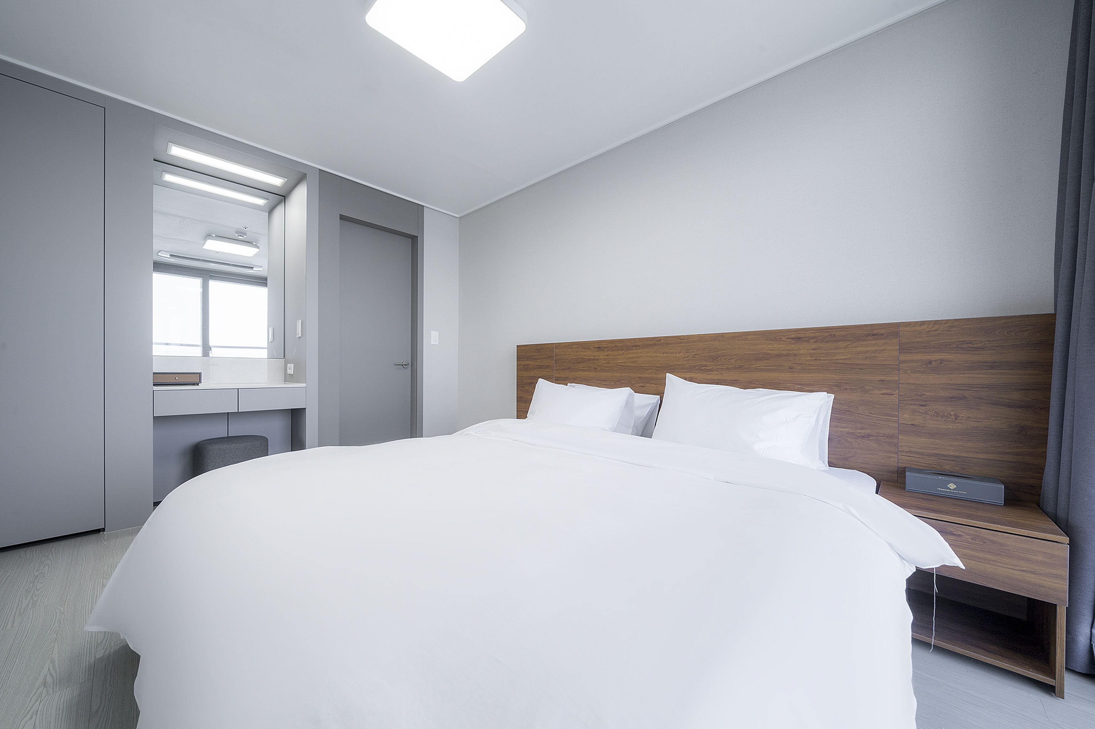 Ein modernes Hotelzimmer mit einem großen Bett, einem Holzkopfstück, einem Nachttisch mit Lampe sowie einem eingebauten Schreibtisch mit Stuhl.