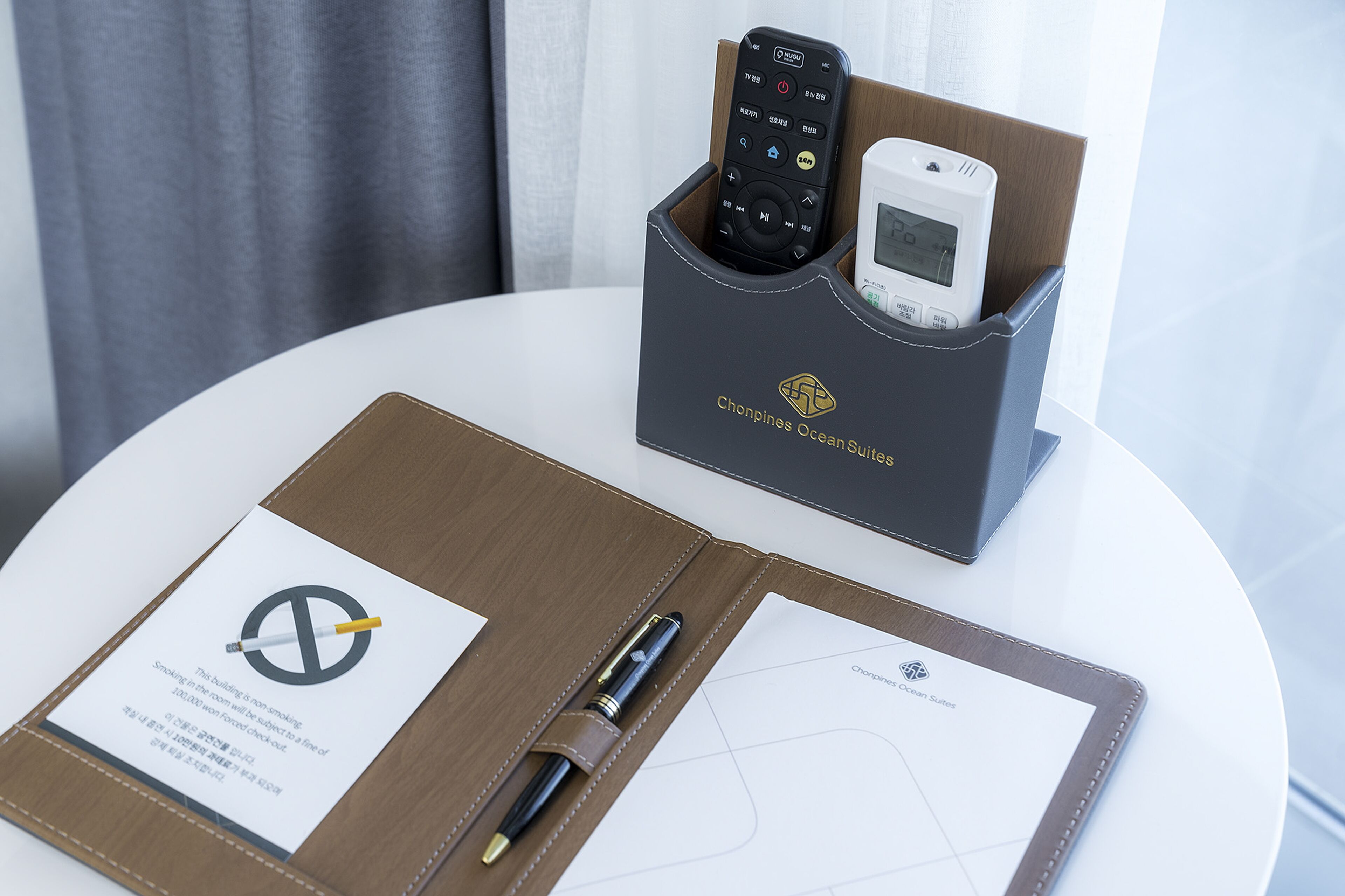 A hotel room with a white table, a brown leather folder, a pen, a remote control, and a white device in a black holder.