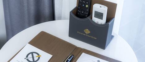 A hotel room with a white table, a brown leather folder, a pen, a remote control, and a white device in a black holder.