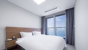 A modern hotel room with a large bed, a wooden headboard, a nightstand, and a view of the sea through a large window. - Gangneung Chonpines Ocean Suites Hotel (Gangneung)