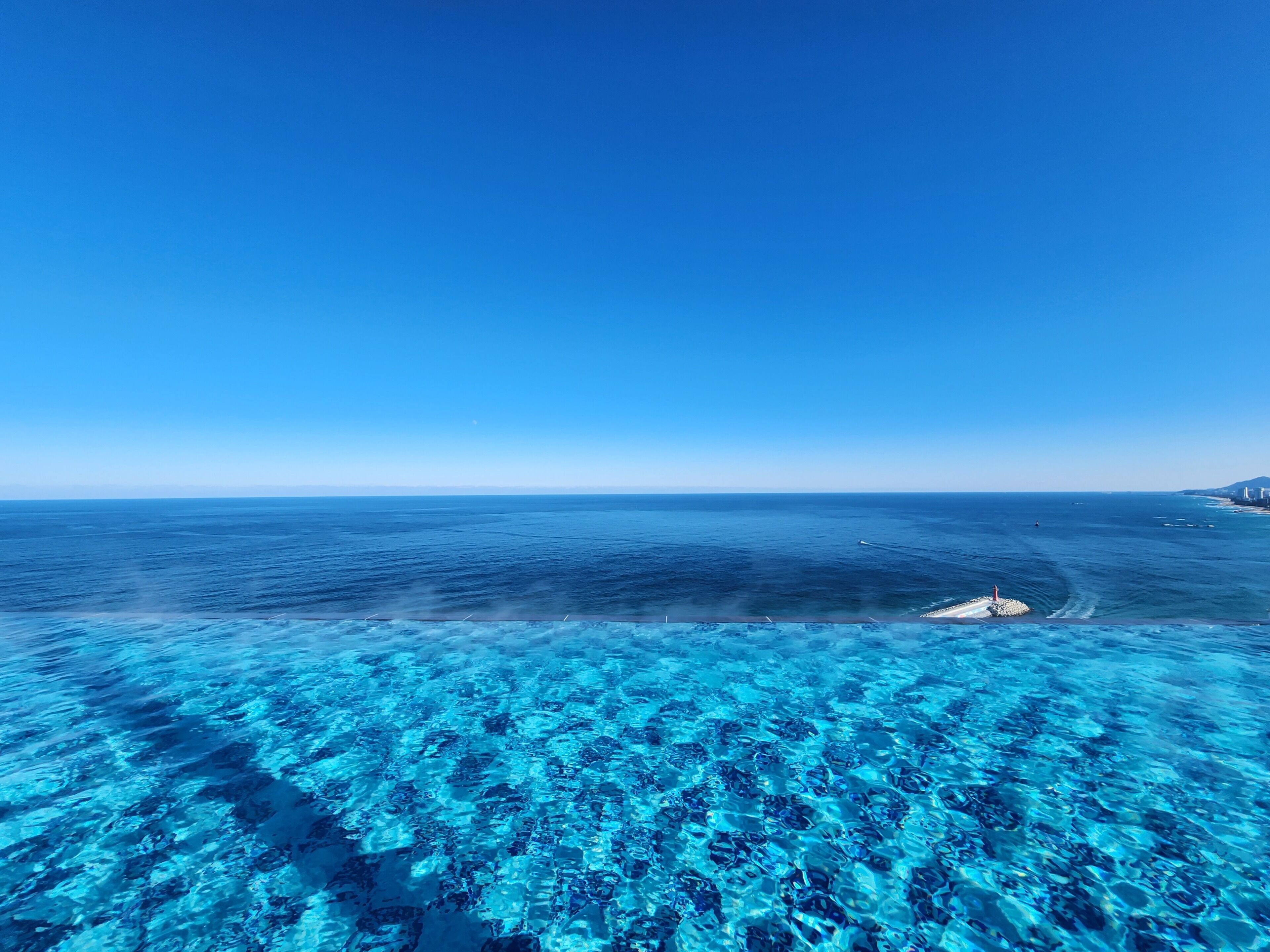 Una vista despejada del océano con un muelle que se adentra en el agua, un barco alejándose de la costa y una línea costera distante con edificios.