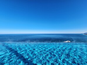 Una vista despejada del océano con un muelle que se adentra en el agua, un barco alejándose de la costa y una línea costera distante con edificios.