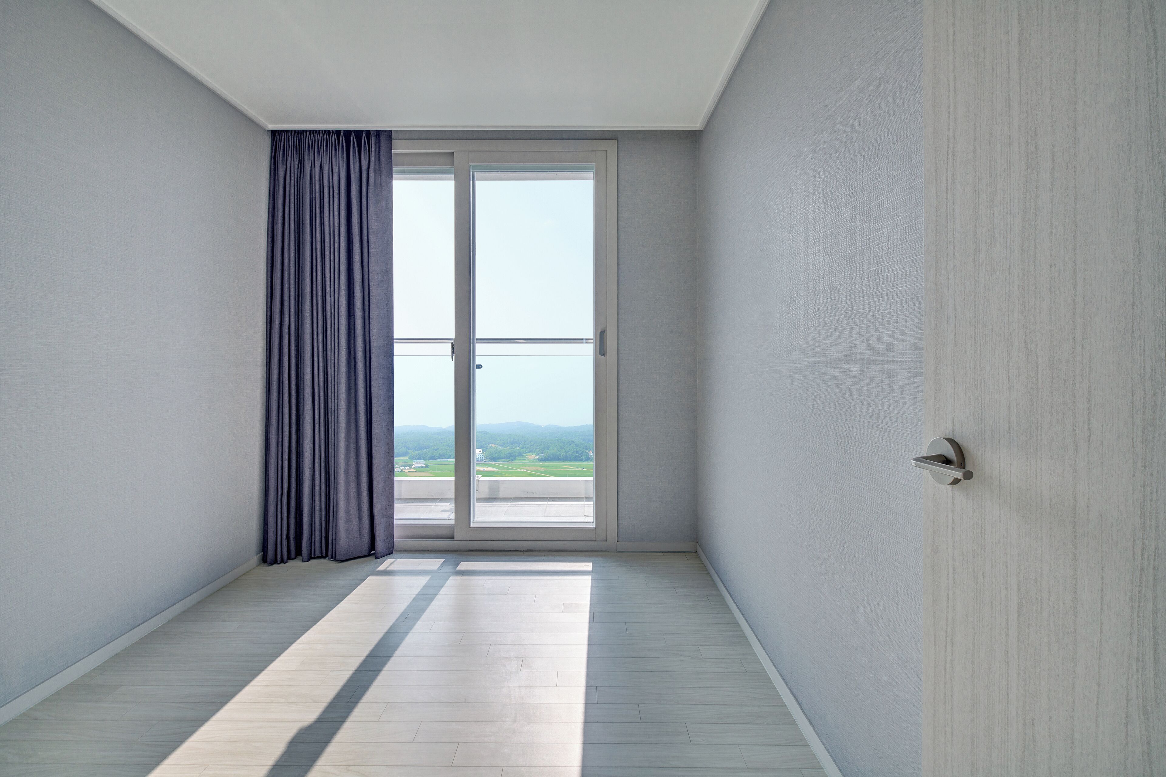 A room with a large sliding glass door leading to a balcony, light-colored wooden flooring, and plain walls.