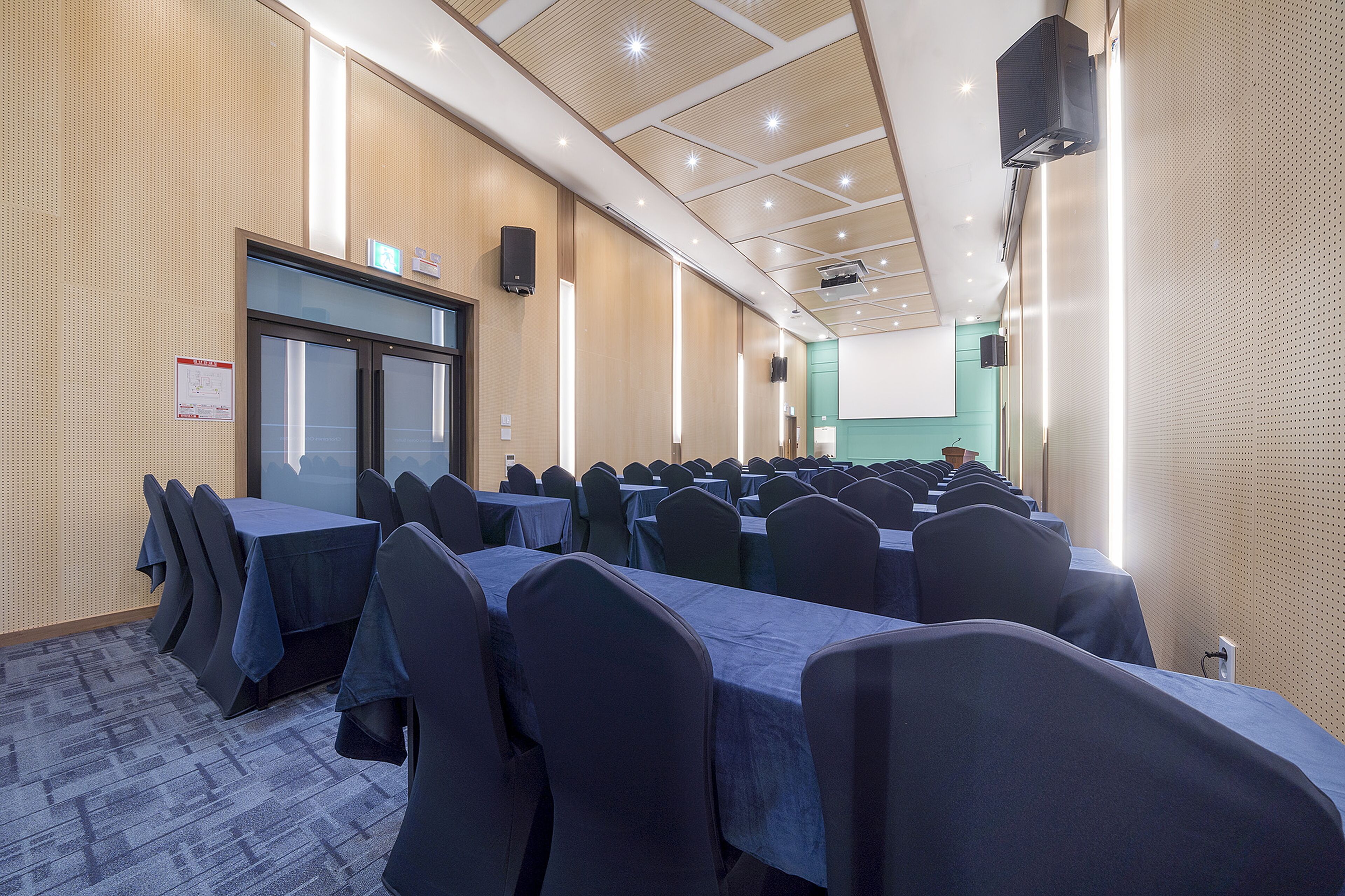 Un salón de conferencias con filas de mesas y sillas, una pantalla de proyector y un altavoz en la pared.