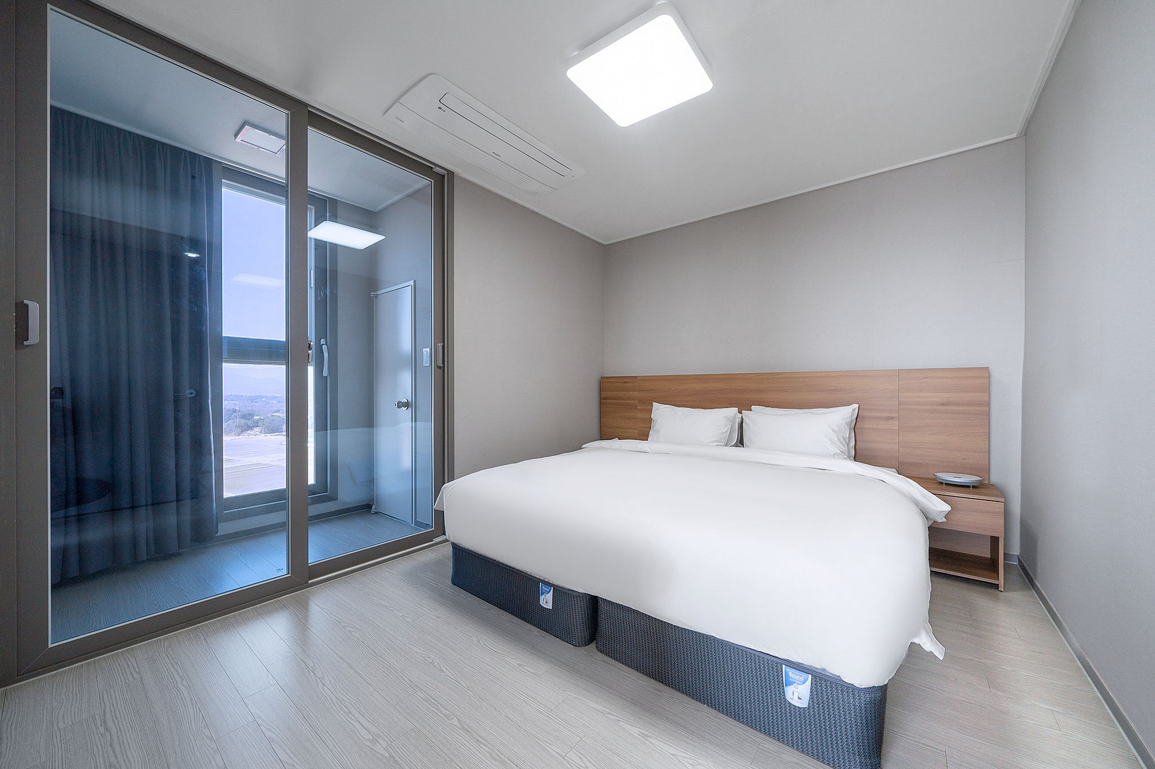 A modern hotel room with a large bed, a wooden headboard, a bedside table, and a sliding glass door leading to a balcony.