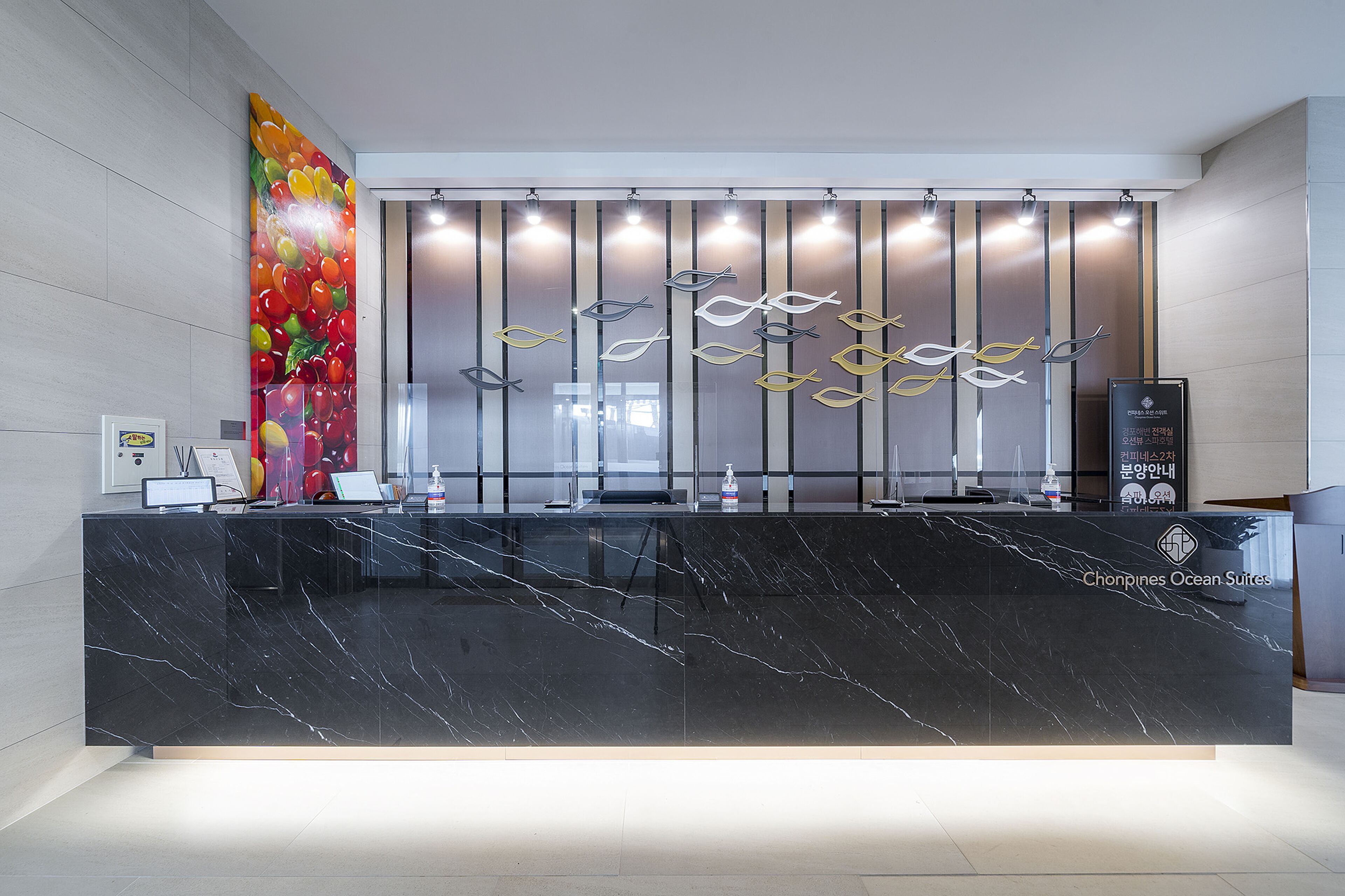 Un comptoir d’accueil moderne, doté d’un plateau en marbre noir, d’une fresque colorée représentant des fruits sur le côté gauche et d’un panneau lumineux vertical au-dessus.