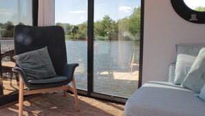 Una habitación con un amplio ventanal que da a una masa de agua, una silla, un sofá y un espejo redondo en la pared.