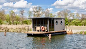 A floating houseboat with a deck, seating area, and a small structure with windows.