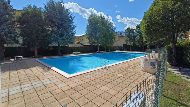 Ein rechteckiger Pool mit einer gefliesten Liegefläche, umgeben von einem Metallzaun und Bäumen.