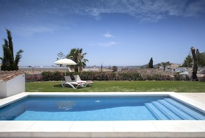 Piscina con sillas de descanso y una sombrilla, rodeada de jardín y vista al mar.