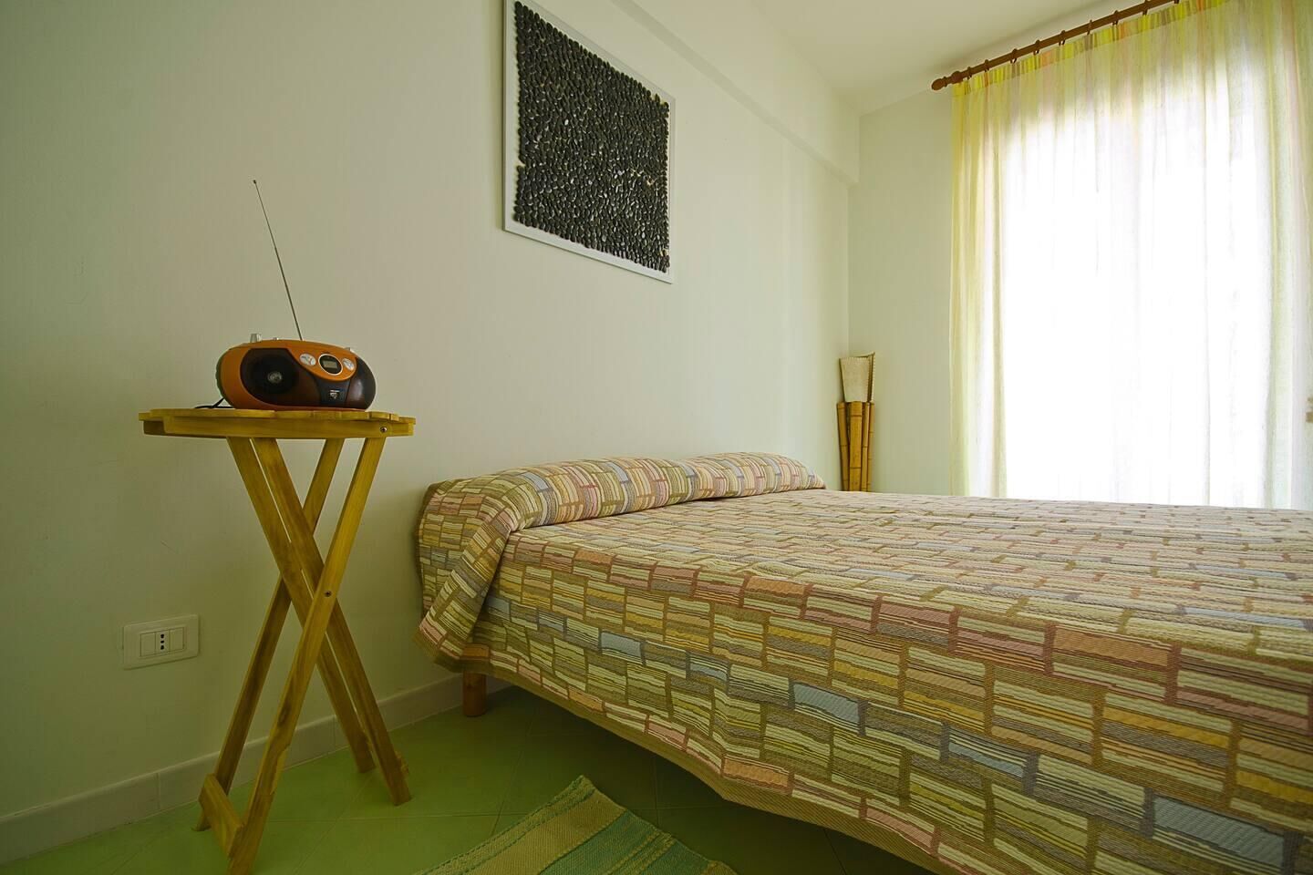 A room with a bed, a radio on a wooden stand, and a window with curtains.