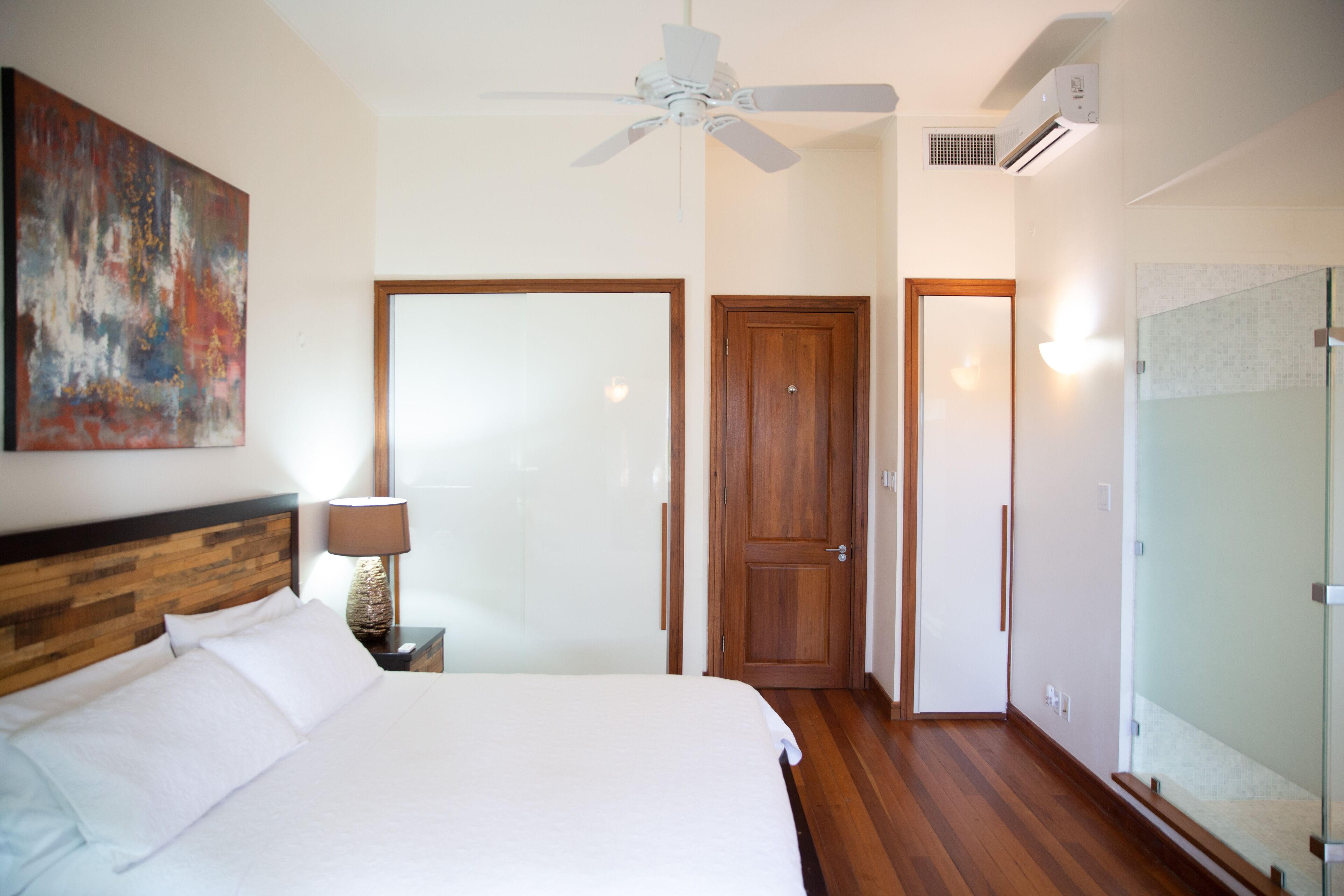 A hotel room with a bed, a ceiling fan, a wooden door, and a glass shower enclosure.