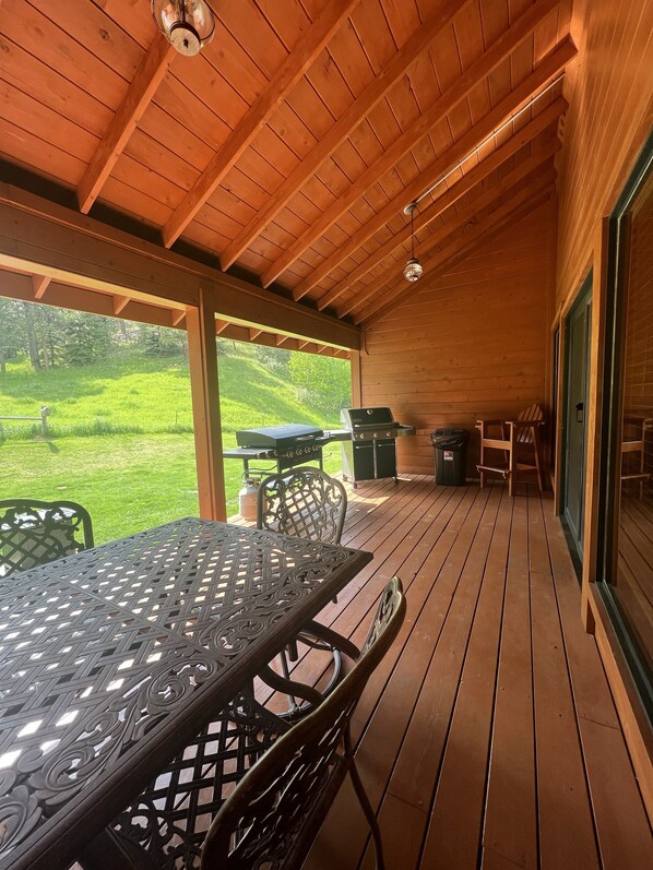A covered outdoor patio with a wooden deck, a metal table with chairs, a grill, and a view of a grassy area.
