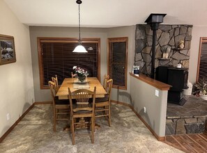Enjoy meals in this cozy dining area with a wooden table and chairs, flanked by large windows and a stone fireplace.