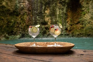 Twee glazen met een drankje en fruitplakjes op een houten dienblad, voor een rotsachtige achtergrond met een wateroppervlak.