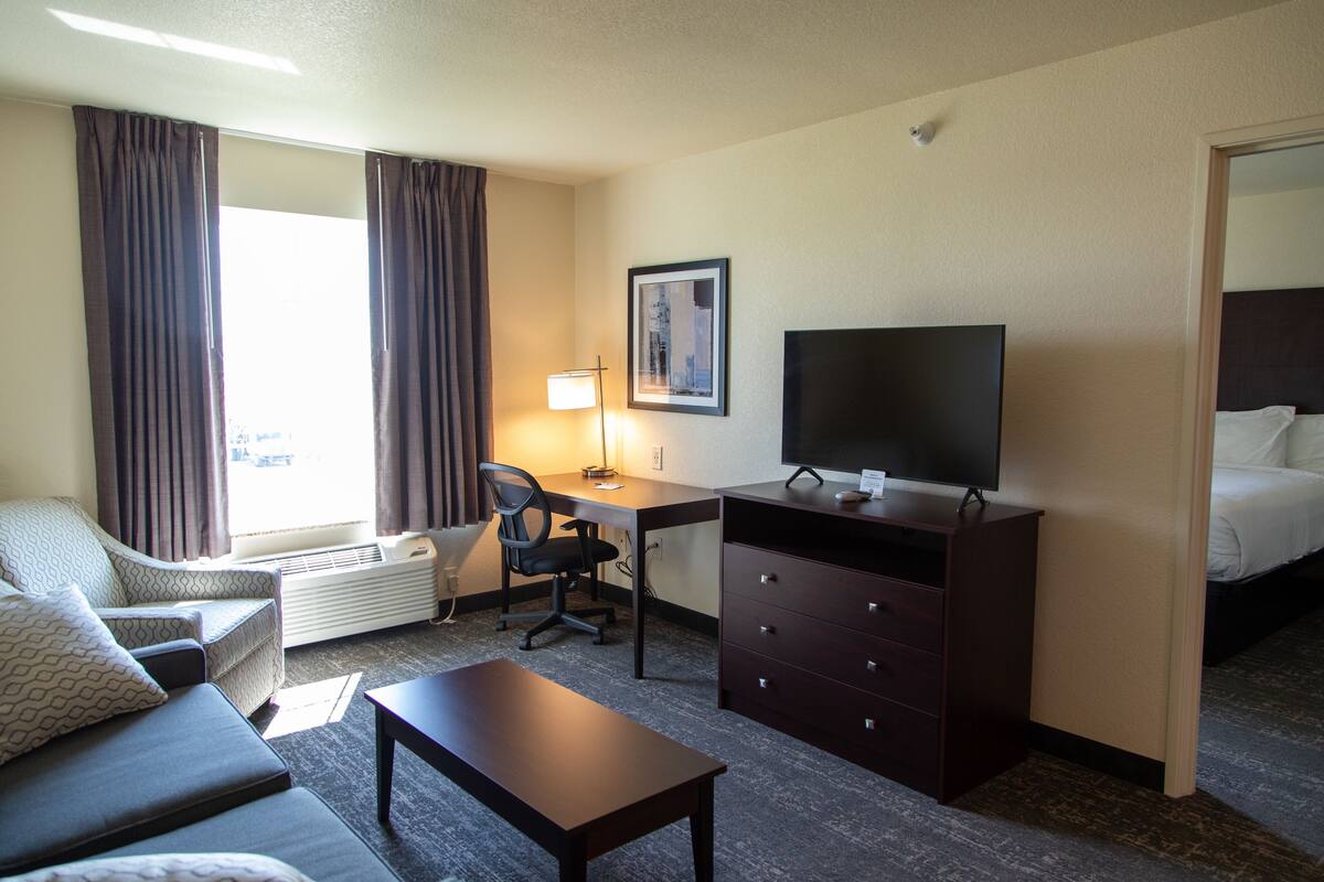A hotel room with a bed, a desk with a chair, a television, a dresser, a sofa, and a window with curtains.
