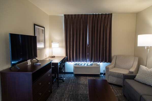 Habitación de hotel con televisión de pantalla plana, escritorio, silla, sofá y ventana grande con cortinas.