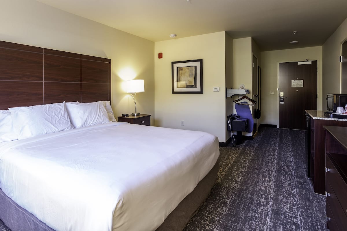 A hotel room with a large bed, a nightstand with a lamp, a dresser, and a small kitchenette.