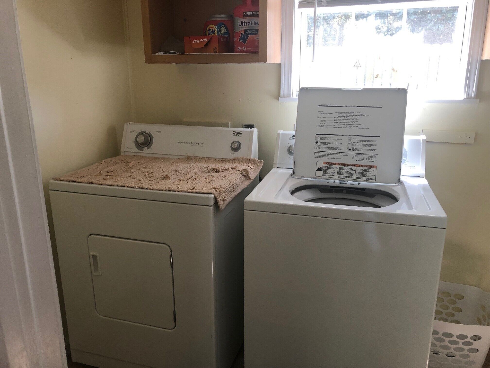 A laundry area with a front-loading washer and dryer, a cabinet above the washer, and a window with blinds.