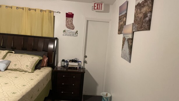 A bedroom with a bed, a nightstand, a door, and wall decorations including a 'Merry Christmas' sign and a landscape painting. - Five minutes (Lauderhill)