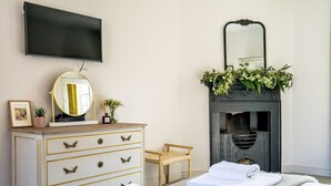Une chambre d’hôtel équipée d’un téléviseur à écran plat, d’un miroir rond, d’une commode avec des poignées dorées, d’une petite chaise en bois, d’une cheminée surmontée d’un miroir et de deux serviettes soigneusement pliées posées sur le lit.