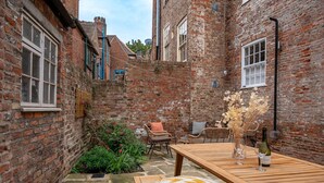 Une cour pavée de briques avec une table en bois, des chaises en osier et un banc. On y trouve des plantes en pot et une bouteille de vin sur la table.