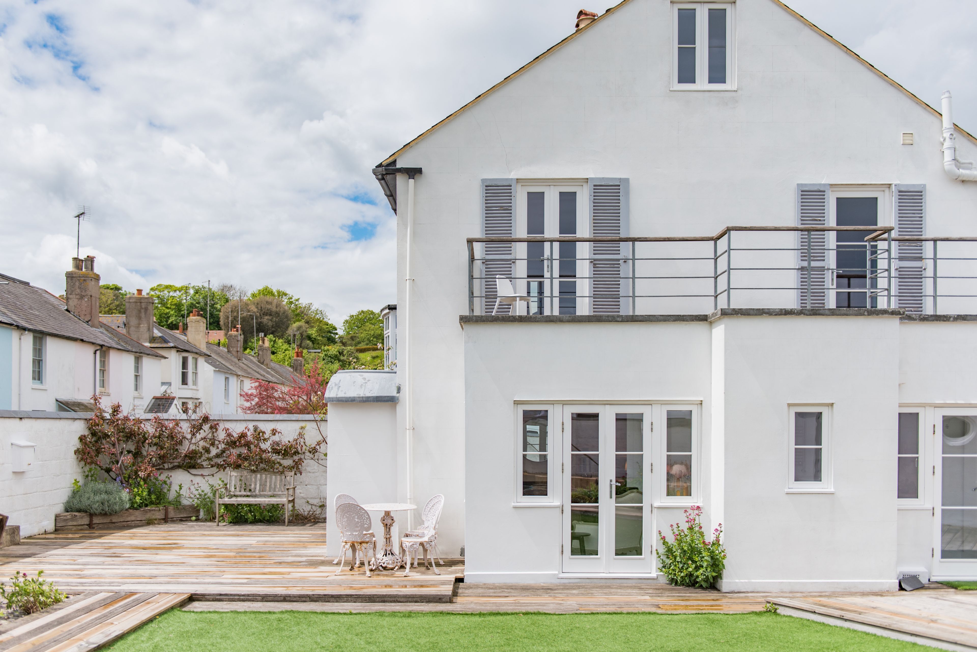 A two-story white house with a balcony, a small patio with a table and chairs, and a well-maintained lawn.