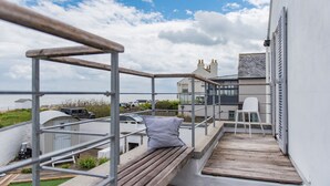 Un balcon avec un banc en bois, une chaise blanche et une rambarde en métal, donnant sur une zone côtière avec des bâtiments et des voitures garées.
