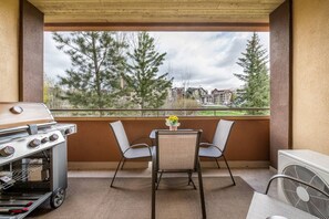 A covered patio with a grill, two chairs, a table with a vase, and a view of trees and buildings.
