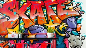 A graffiti wall with the word "SKATE" in large letters, a table with two potted plants, and red chairs. - Rucksack Inn Temple Street (Singapore)