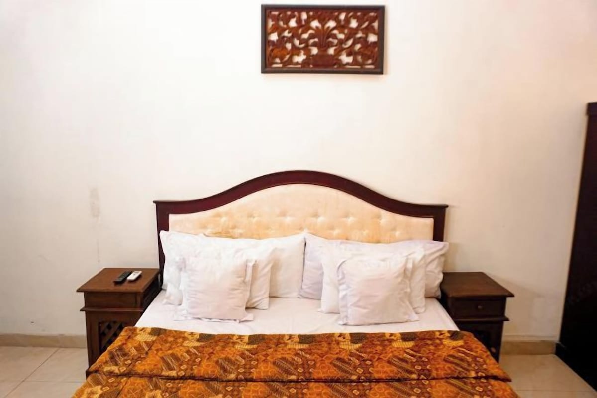 Een netjes opgemaakt bed met een beddekleed met patroon, twee nachtkastjes waarvan er één een afstandsbediening heeft, en een decoratief wandobject boven het bed.