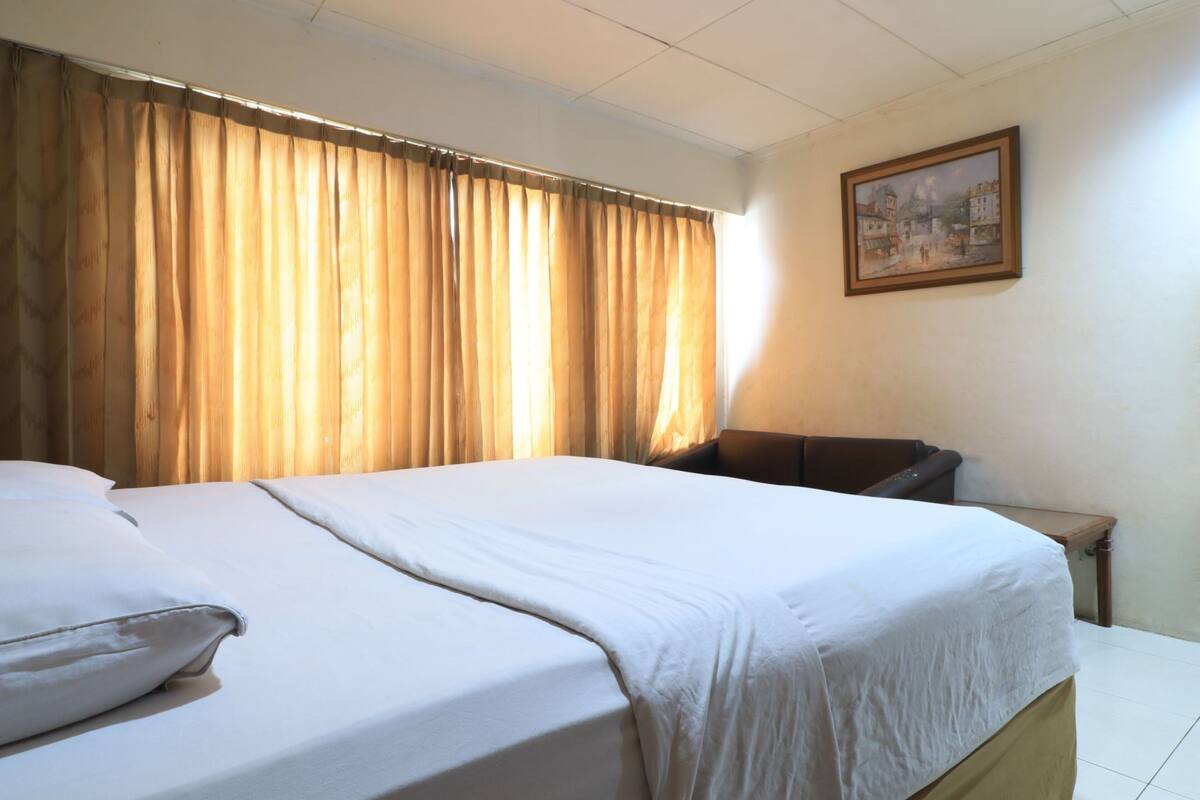 Une chambre d’hôtel avec un grand lit, une tête de lit en bois, un fauteuil en cuir marron, une petite table et un tableau accroché au mur.