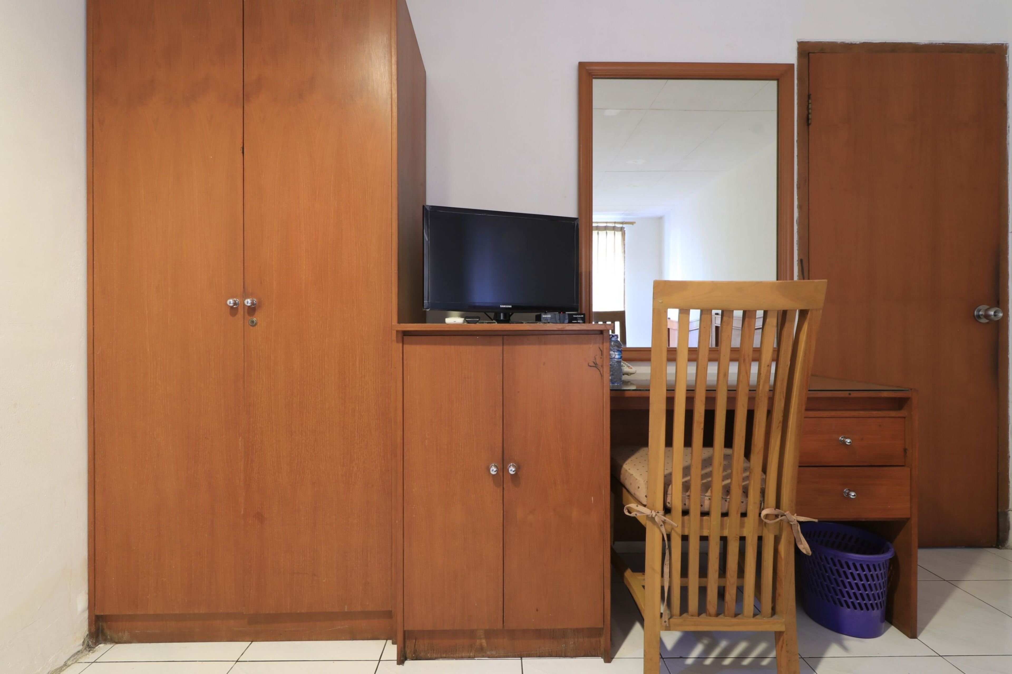 Un dormitorio con un armario de madera, una cama con cabecera de madera, una mesita de noche pequeña de madera y un televisor sobre una estantería.